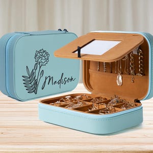 K&ouml;nnte beinhalten: Hellblaue Schmuckschatulle mit floralem Design und dem Schriftzug "Madison". Die Box ist ge&ouml;ffnet und zeigt F&auml;cher f&uuml;r Ringe und Halsketten. Der Innenraum ist beige, auf einer Holzoberfl&auml;che.