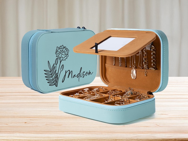 K&ouml;nnte beinhalten: Hellblaue Schmuckschatulle mit floralem Design und dem Schriftzug "Madison". Die Box ist ge&ouml;ffnet und zeigt F&auml;cher f&uuml;r Ringe und Halsketten. Der Innenraum ist beige, auf einer Holzoberfl&auml;che.