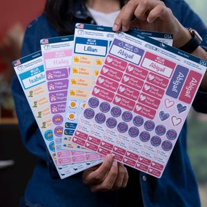 May include: A person holds several sheets of name labels with various designs. The labels feature names like Abigail, Lillian, and Isabella, along with decorative elements such as hearts, stars, and dinosaurs. The labels are in pink, blue, and purple.