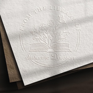May include: Embossed stationery with a floral and butterfly design. The text reads "FROM THE LIBRARY OF" above an open book with flowers and a butterfly, and "Susanne Dawson" below. The stationery is on a stack of paper.