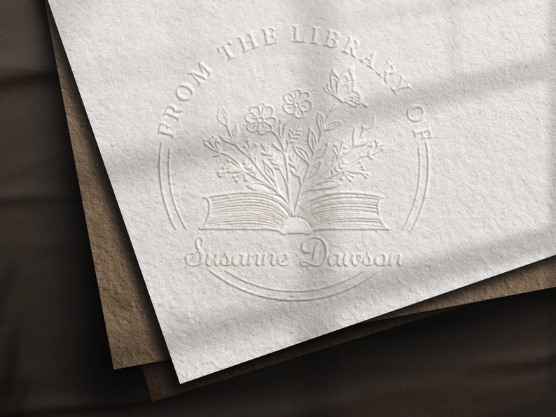 May include: Embossed stationery with a floral and butterfly design. The text reads "FROM THE LIBRARY OF" above an open book with flowers and a butterfly, and "Susanne Dawson" below. The stationery is on a stack of paper.