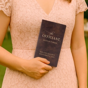 May include: A dark brown wedding program held by a person wearing a pink lace dress. The program reads "the Officiant" with the names "Hannah Parker", "Miranda & Kaden", and the date "September 6, 2025".