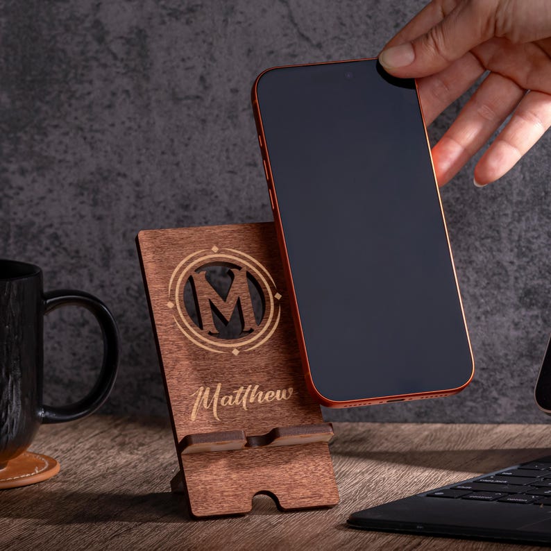 May include: A wooden phone stand with the letter "M" inside a circle, and the name "Matthew" engraved below. A red-framed smartphone is placed on the stand. A black mug and a laptop are also visible.