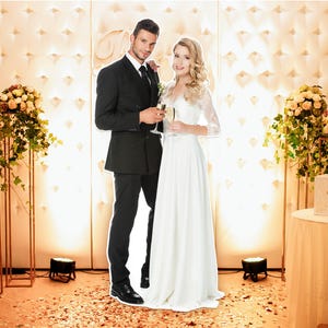 May include: A couple in formal attire, holding champagne flutes, stands in front of a decorated backdrop. The woman wears a white wedding dress with lace sleeves. The man is in a black suit. Floral arrangements and a table with a decorative box are visible.