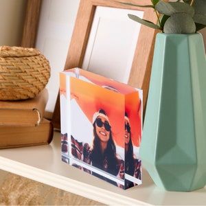 May include: A clear acrylic photo block displaying a vibrant image of two people wearing sunglasses. The block is rectangular and stands upright on a white shelf, next to a light green geometric vase and a woven basket. A wooden picture frame is in the background.