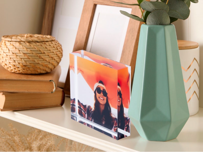 May include: A clear acrylic photo block displaying a vibrant image of two people wearing sunglasses. The block is rectangular and stands upright on a white shelf, next to a light green geometric vase and a woven basket. A wooden picture frame is in the background.
