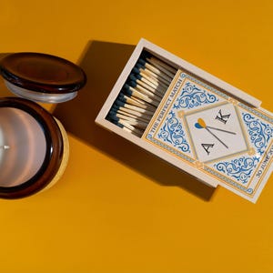 May include: A brown glass candle jar with a white candle and a lid, next to a matchbox. The matchbox has a decorative design with the text "THE PERFECT MATCH" and initials "A K" with the date "30 JUNE 2025".
