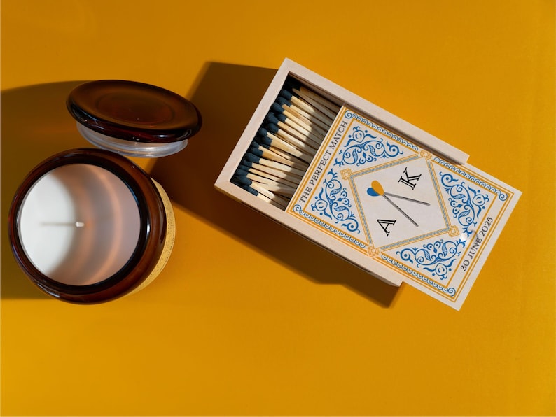 May include: A brown glass candle jar with a white candle and a lid, next to a matchbox. The matchbox has a decorative design with the text "THE PERFECT MATCH" and initials "A K" with the date "30 JUNE 2025".