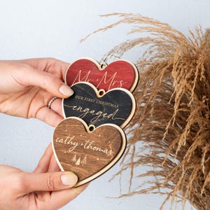 May include: Three wooden heart-shaped ornaments with the text "Mr. & Mrs." on the top ornament, "Our First Christmas Engaged" on the middle ornament, and "cathy + thomas" on the bottom ornament.