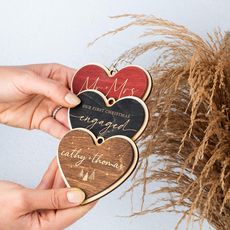 May include: Three wooden heart-shaped ornaments with the text "Mr. & Mrs." on the top ornament, "Our First Christmas Engaged" on the middle ornament, and "cathy + thomas" on the bottom ornament.
