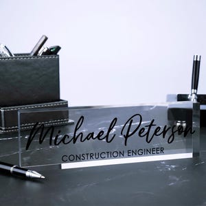 May include: A clear acrylic desk nameplate with black text that reads "Michael Peterson, Construction Engineer".