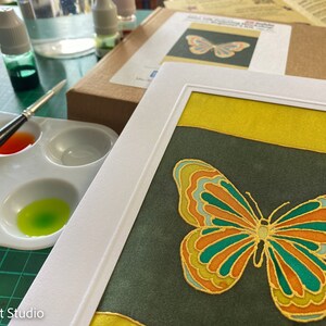 May include: A framed artwork featuring a colorful butterfly with green, orange, and gold accents on a dark green background. The butterfly is surrounded by a yellow border.