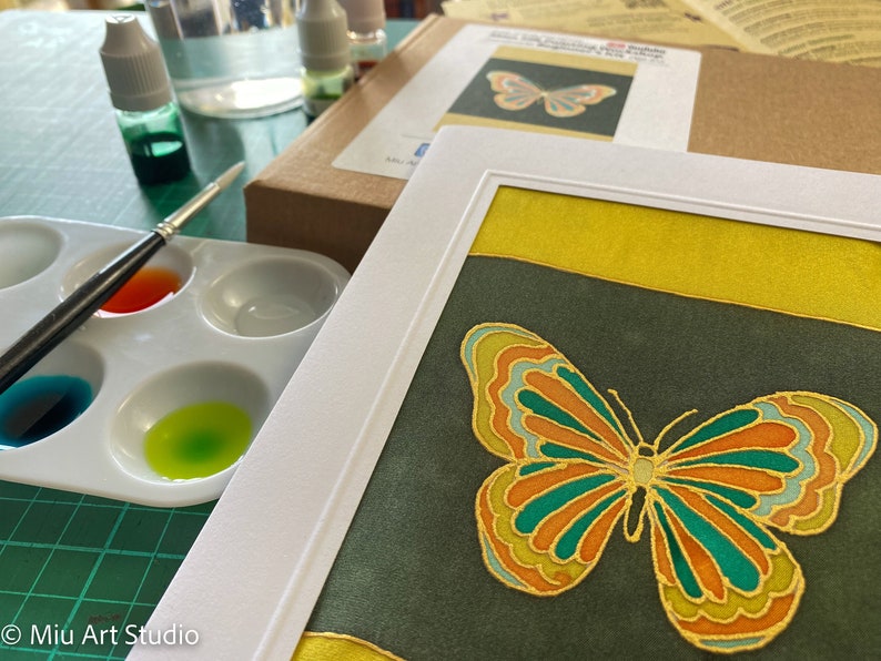 May include: A framed artwork featuring a colorful butterfly with green, orange, and gold accents on a dark green background. The butterfly is surrounded by a yellow border.