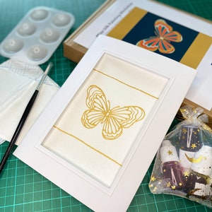 May include: A gold butterfly painting kit with a white frame, a paintbrush, a palette, and a bag of paint.