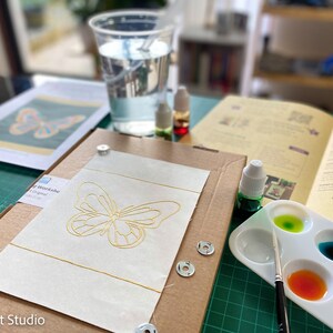 May include: A butterfly embroidery kit with a gold butterfly design on white fabric, a small paint palette with green, blue, and orange paint, and a paintbrush.