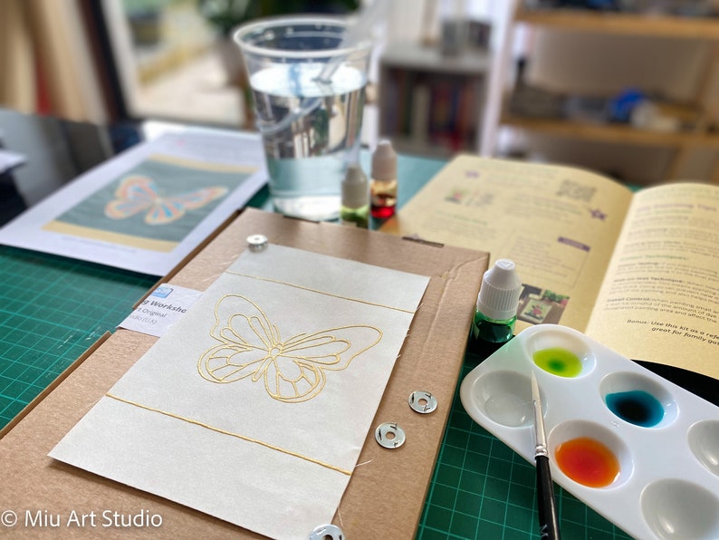 May include: A butterfly embroidery kit with a gold butterfly design on white fabric, a small paint palette with green, blue, and orange paint, and a paintbrush.