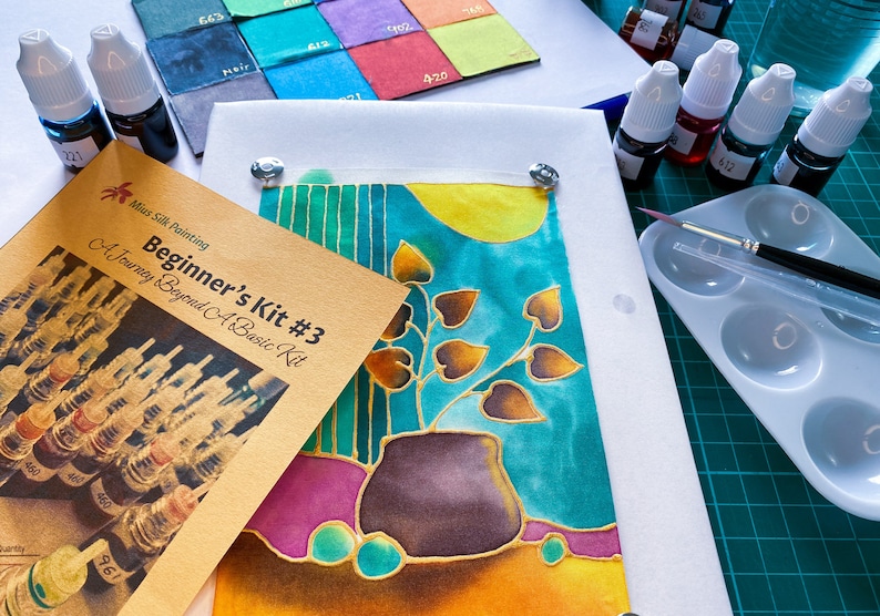 May include: A beginner's silk painting kit with a guidebook, silk fabric, paints, and tools. The silk fabric features a painted design of a plant in a pot with leaves in shades of brown and gold against a turquoise background.