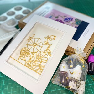 Pode incluir: Uma moldura branca com um passe-partout branco e um desenho floral dourado sobre fundo branco. A moldura est&aacute; sobre uma esteira de corte verde com tr&ecirc;s frascos de tinta e uma pequena bolsa de glitter dourado.