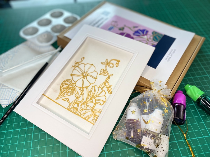 Pode incluir: Uma moldura branca com um passe-partout branco e um desenho floral dourado sobre fundo branco. A moldura est&aacute; sobre uma esteira de corte verde com tr&ecirc;s frascos de tinta e uma pequena bolsa de glitter dourado.