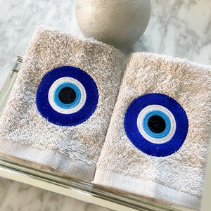 May include: Two beige hand towels with embroidered blue and white evil eye designs. The towels are displayed on a mirrored tray, with a silver sphere candle in the background. The towels have a textured appearance.