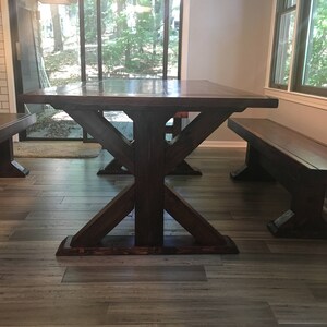 May include: A dark brown wooden table with a farmhouse style X-shaped base and two matching benches. The table and benches are in a room with hardwood floors and a window with a view of trees.