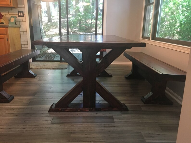 May include: A dark brown wooden table with a farmhouse style X-shaped base and two matching benches. The table and benches are in a room with hardwood floors and a window with a view of trees.