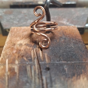 May include: A handcrafted copper wire ring with a unique, swirling design. The ring features multiple loops and curves, creating an elegant and artistic look. The ring is shown on a wooden surface with a metal tool in the background.