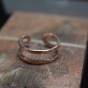 May include: A copper cuff bracelet with a double-band design. The open-ended bracelet has a textured surface and sits on a wooden surface. The warm, reddish-brown metal contrasts with the wood grain.