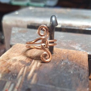 May include: A handcrafted copper ring with a unique, swirling design. The ring features two bands and decorative spirals, showcasing a delicate, artistic style. The ring is displayed on a wooden surface.