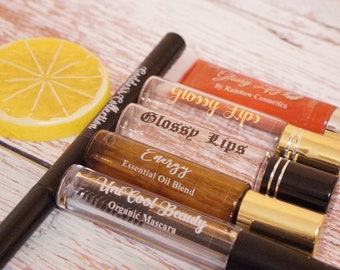 May include: An assortment of beauty products including lip gloss, mascara, and essential oil blend. The lip gloss tubes are labeled "Glossy Lips" and the mascara is labeled "Uni Cool Beauty". A yellow lemon-shaped soap is also present.