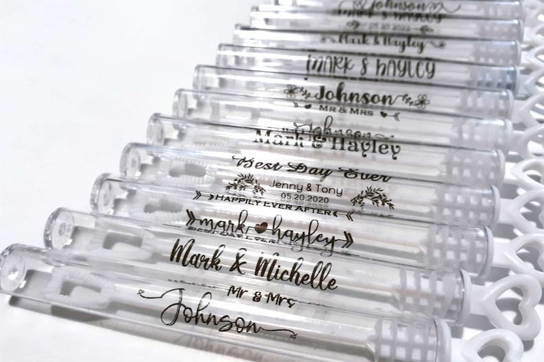 May include: Clear plastic bubble wands with white heart-shaped handles. Each wand is personalized with black text, including names, dates, and phrases like "Best Day Ever" and "Happily Ever After." Ideal for weddings or celebrations.