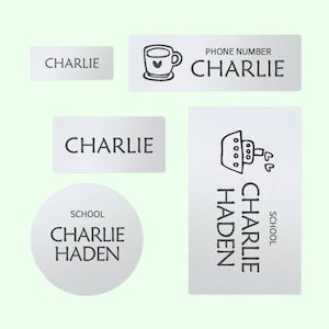 May include: A collection of personalized name tags and labels. The labels are white with black text, featuring the name "CHARLIE" on several. One label includes a coffee cup graphic, and another says "CHARLIE HADEN SCHOOL".
