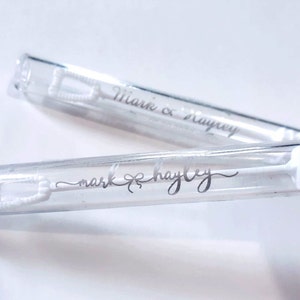 May include: Two clear bubble wands with white bubble solution and wands. The wands have the names "Mark & Hayley" printed in silver script. The bubble wands are a popular item for weddings and other celebrations.
