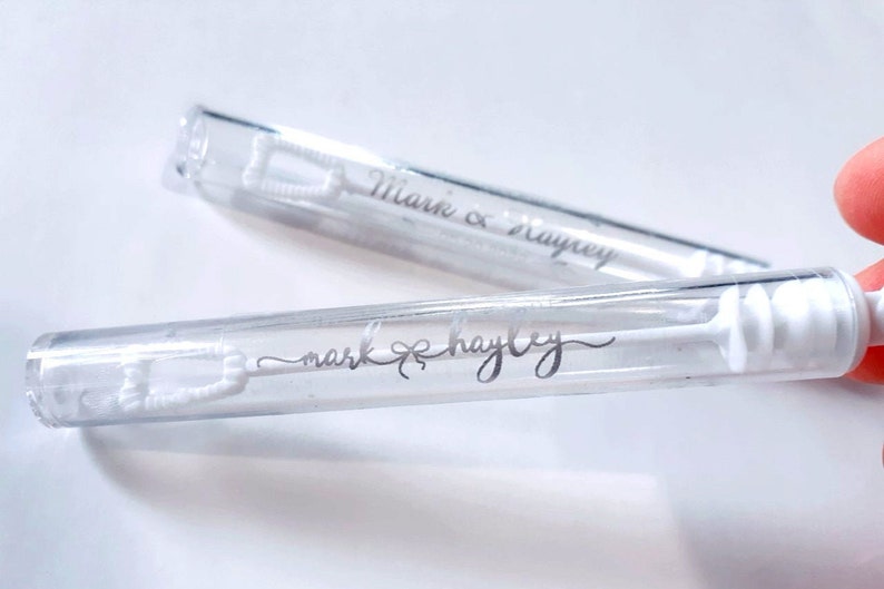 May include: Two clear bubble wands with white bubble solution and wands. The wands have the names "Mark & Hayley" printed in silver script. The bubble wands are a popular item for weddings and other celebrations.