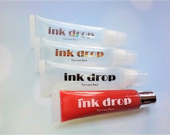 May include: Four clear lip gloss tubes with the text "ink drop" and "Tanned Red" printed on the labels. One tube is filled with a red lip gloss.