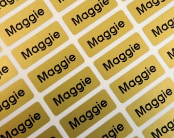 May include: A sheet of rectangular gold-colored adhesive labels arranged in a grid pattern. Each individual label features the name Maggie printed in a bold, black, sans-serif font. The labels are uniform in size and shape, designed for identification.