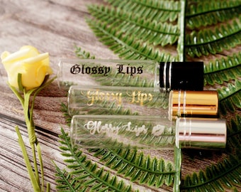 May include: Three clear glass bottles with gold lettering that reads "Glossy Lips" and different colored roll-on tops. The bottles are on a wooden surface with green foliage.