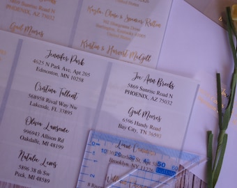 May include: A close-up of a set of address labels with names and addresses printed in gold lettering on a white background. The labels are arranged in a grid pattern and are surrounded by a ruler and a small flower.