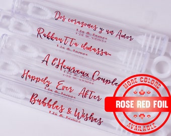 May include: A set of clear plastic bubble wands with various wedding-themed messages printed in rose red foil. The messages include "Dos corazones y un Amor", "Rakkautta ilmassa", "A l'Heureux Couple", "Happily Ever After", and "Bubbles & Wishes". The image also includes a red circle with the text "MORE COLORS ROSE RED FOIL AVAILABLE".