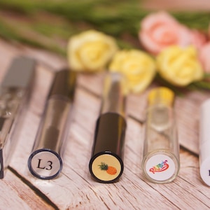 May include: Five small glass bottles with colorful labels. The labels have different designs, including a pineapple, a kiwi, and the word "L3".