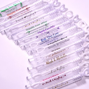 May include: Clear plastic bubble wands with heart-shaped handles. Each wand is labeled with wedding-themed text in various colors, including "Happily Ever After" and names like "Mark & Hayley".