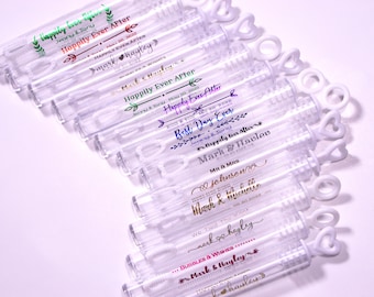 May include: A collection of clear plastic bubble wands with personalized messages for a wedding. The wands have a heart-shaped handle and are decorated with gold, silver, and colorful text. The text includes the names "Mark & Hayley" and the phrase "Happily Ever After".