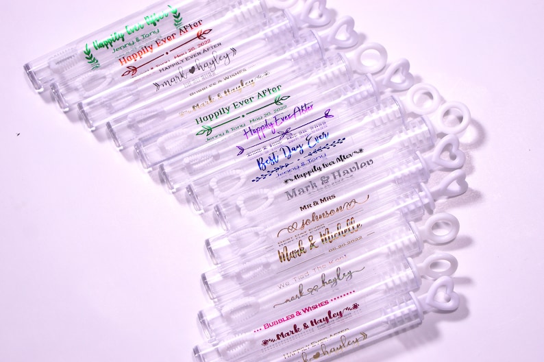 May include: Clear plastic bubble wands with heart-shaped handles. Each wand is labeled with wedding-themed text in various colors, including "Happily Ever After" and names like "Mark & Hayley".
