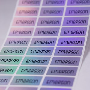 May include: A sheet of iridescent holographic stickers with the name "Emerson" printed on each sticker. The stickers are arranged in rows and columns, with the name "Emerson" repeated on each sticker.
