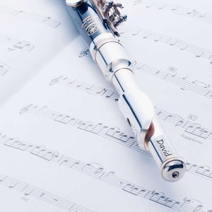 May include: A silver and black Yamaha piccolo flute rests on a sheet of musical notes. The flute has the name "David" engraved on it. The background is a soft, light blue.