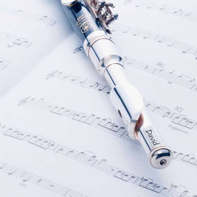 May include: A silver and black Yamaha piccolo flute rests on a sheet of musical notes. The flute has the name "David" engraved on it. The background is a soft, light blue.