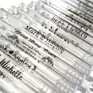 May include: Clear plastic bubble wands with personalized black text. The wands are arranged in rows and feature names, dates, and phrases like "Best Day Ever" and "Happily Ever After." The wands are on a white surface.