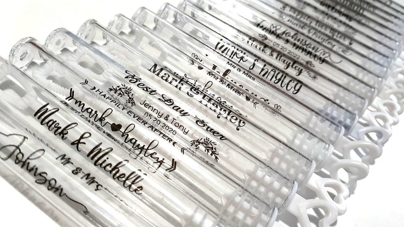 May include: Clear plastic bubble wands with personalized black text. The wands are arranged in rows and feature names, dates, and phrases like "Best Day Ever" and "Happily Ever After." The wands are on a white surface.