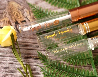 May include: Three clear glass bottles with gold and silver caps. The bottles contain lip gloss and have labels with the text "Angel Lips", "Angel Beauty Lip Gloss", and "Bossy Gloss By R.L. Cosmetics".