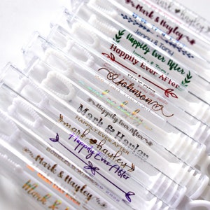 May include: Clear plastic bubble wands with white handles. Each wand has personalized text in various colors, including "Mark & Hayley" and "Happily Ever After." Ideal for weddings or celebrations.